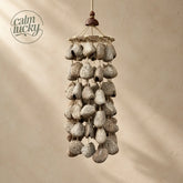 Large handmade driftwood-grey textured seed pod wind chime, dense multi-tier cylindrical cascade of weathered natural shells on a woven ring, calming forest rain sound decor on beige background