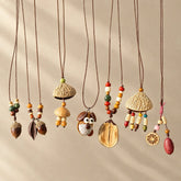 Nine handmade natural seed necklaces in a row, including carved acorns, nut bell pendants, beaded strands and citrus slice charms, on adjustable brown cords — boho nature jewelry collection on beige background
