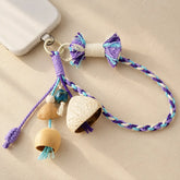 Handmade indigo-purple and sky-blue woven seed bell phone charm with a white seed cap pendant, colorful tassel bow and braided wrist strap attached to a mobile phone keyring, boho nature accessory on beige background