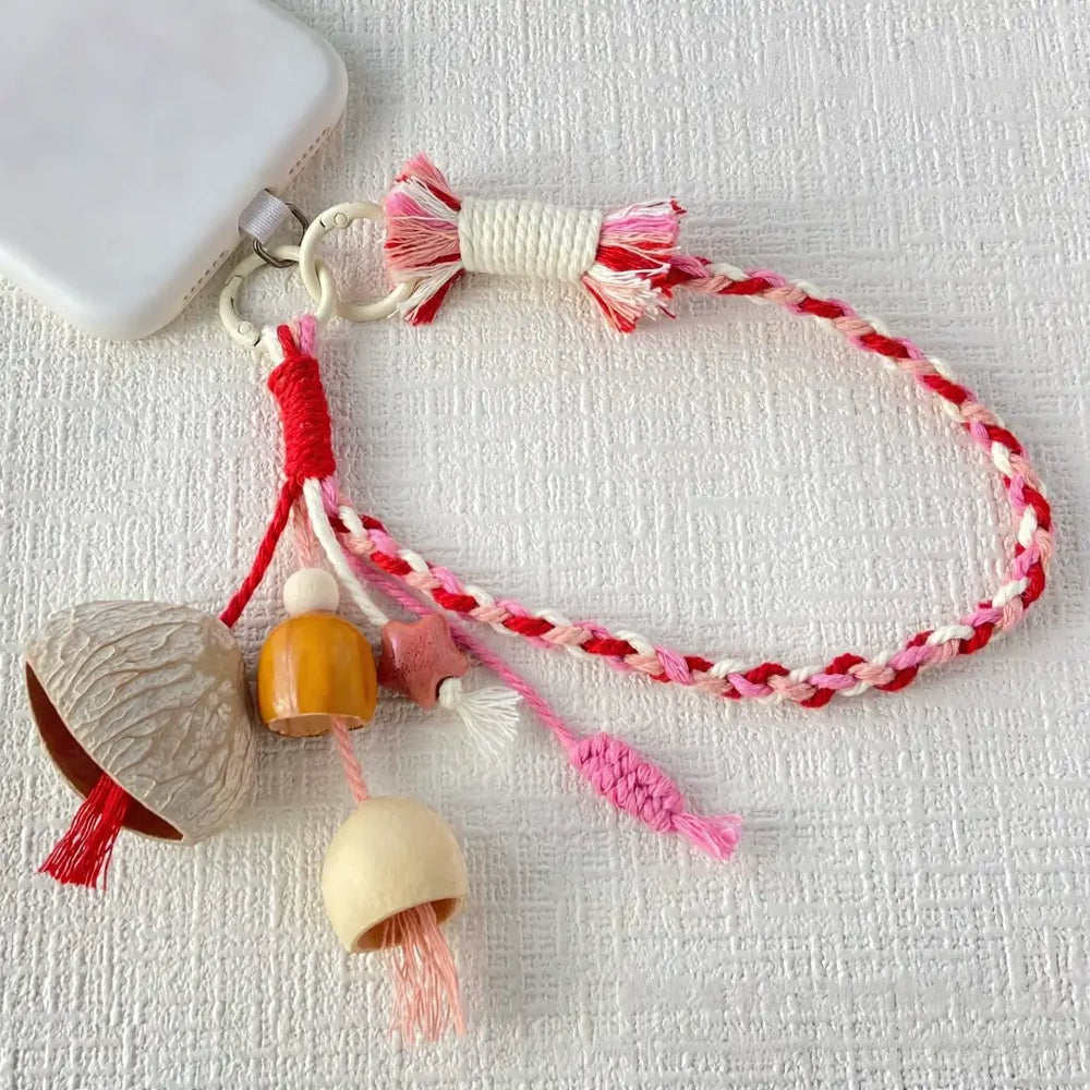 Rose Hip seed bell phone charm — red and pink hand-braided wrist strap with ivory nut shell bell and wooden beads, clipped to a phone